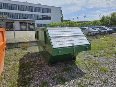 Frontansicht einer grünen 7 m³-Absetzmulde mit beidseitig klappbarem Riffelblechdeckel, abgestellt auf Schotterfläche vor Bürogebäude.