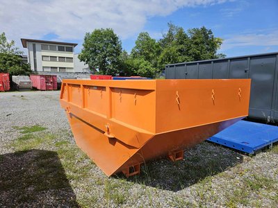 Rückansicht einer stapelbaren, offenen 10 m³-Absetzmulde (Sperrgutmulde) in Orange, stehend auf Kiesfläche vor Grünanlage und Bürogebäude.