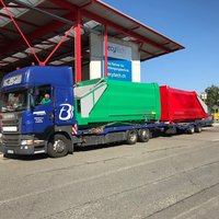 Zwei Abrollpresscontainer (grün und rot) auf blauem LKW unter Ladehalle – effizienter Doppeltransport für Mulden- und Abfallverdichtung.